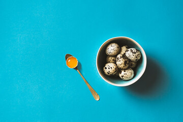 Quail eggs in a bowl on blue background. one cracked egg with yellow yolk in spoon.