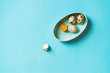 Quail eggs in a bowl on blue background. One cracked egg with yellow yolk and egg shell
