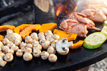 Close-up detail view of many tasty raw marinated quails and vegetables and mushrooms grilled cooked at round steel iron firepit hearth table surface brazier with burning firewoods. Barbecue yard home