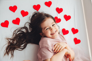 Valentine's Day, a little girl lying at home on a white bed among hearts, smiling and laughing with happiness, congratulating on the holiday