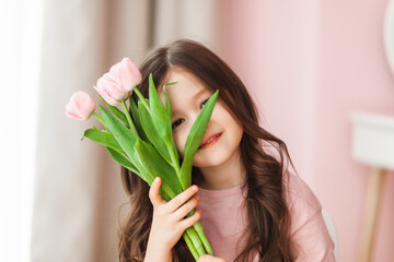 Naklejka premium Portrait of a little girl with long dark hair close-up. The baby hugs a bouquet of fresh, delicate pink tulips. A gift for the holiday, spring time.