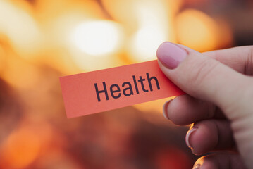Female hand holding red sheet of paper with printed word "health" on it, burning fire on background. Concept of welfare, well-being, soundness