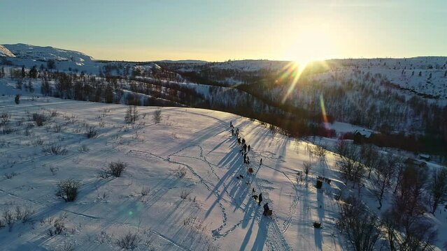 Aerial parallax shot of people standing in long queue with their huskies on top of a mountain landscape covered with snow and beautiful reflection of sunlight falling on the mountains