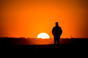 Fototapeta premium Silhouette of a man with a coat and his hands in his pockets, watching the sunset