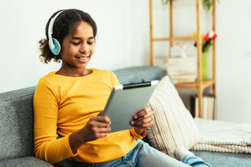 Happy African young girl using digital tablet technology sitting on couch at home.