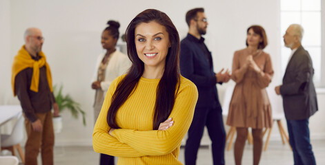 Portrait of successful female employee and business professional at work. Happy beautiful young brunette woman in yellow jumper standing in office with her arms crossed, looking at camera and smiling