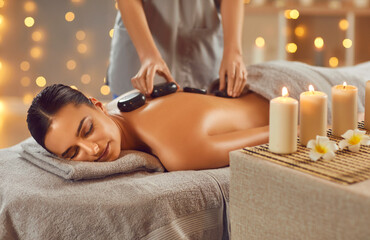 Woman enjoying exotic hot stones spa massage. Relaxed young woman lying on a spa bed while the masseuse is putting hot stones on her back. Spa treatment concept