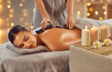 Wall murals Spa Woman enjoying exotic hot stones spa massage. Relaxed young woman lying on a spa bed while the masseuse is putting hot stones on her back. Spa treatment concept