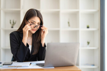 Photo of a beautiful woman holding eyeglasses while sitting at the working desk surrounded by a computer laptop, paperwork, calculator, coffee cup at office.