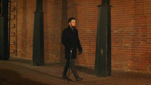 Bearded Guy In Coat Walks Leaning On Cane Past Brick Building At Night. Dude, Dandy Walking At Emptied Urban Street At Late Evening. Tired Caucasian Man Returns Home After Work On Foot