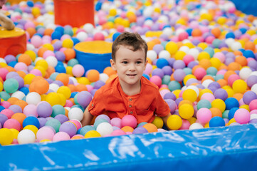 Fototapeta na wymiar Portrait of a happy little boy jumping merrily into a dry pool with colorful balloons. A toddler boy is playing and having a good time at the children's entertainment center.