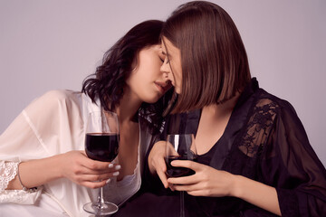 Valentine's day concept. Portrait of two lesbians in satin negligee holding glasses of red wine while sitting on a sofa, intimate moments of private life. Two girls are relaxing on a couch at home.