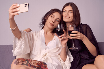 Valentine's day concept. Portrait of two smiling lesbian girls in satin negligee taking selfies. Girls are holding glasses of red wine while sitting on a sofa. Cropped shot.