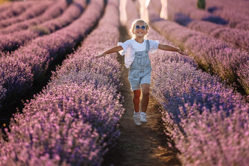 Adorable happy child girl runs raising her hands like flying plane in lavender field on summer warm day. Hyperactive smiling little kid in sunglasses dreams of flying in nature. Children's fantasies.