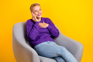 Portrait of excited cheerful girl sit chair speak telephone hand touch chest laughing isolated on yellow color background