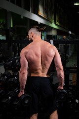Fototapeta na wymiar A brutal muscular man with a beard and a perfect body stands with his back and holds dumbbells in his hands in the gym. Fitness, beauty