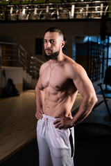 Fototapeta na wymiar A brutal muscular man with a beard and a perfect body poses in the gym in white pants. Fitness, beauty