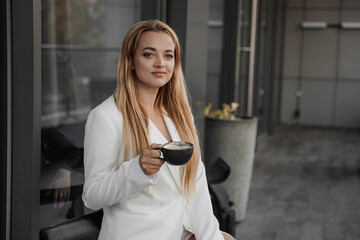 Fototapeta na wymiar Blond stylish woman at oversized white jacket drinking aroma coffee and relax on the modern city background