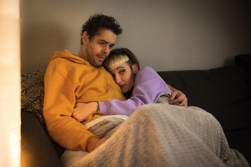 Biracial couple stretched out on the sofa covered with the blanket scared watching a horror movie on TV