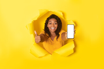 Joyful african american lady holding smartphone with blank screen and showing thumb up posing...