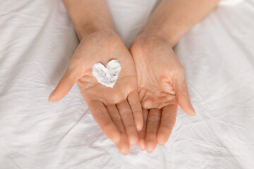 Caucasian millennial lady applying heart shaped cream on hands, enjoy good morning in bedroom, top view
