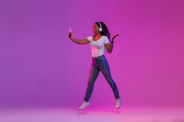 Joyful Black Woman In Neon Light Jumping With Smartphone In Hands