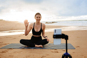 Plakat Online training. Sporty woman yoga coach recording video online training, sitting on fitness mat on the beach by seaside