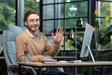 Portrait of a young businessman, designer, programmer in a busy office sitting at a table with a computer, smiling at the camera, waving, greeting, introducing himself.