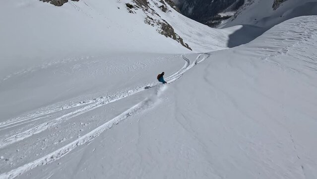 Male snowboarding in blue overalls clothing extreme heliski freeride from mountain fresh snow summit red avalanche backpack aerial view. FPV sport drone dynamic shot man speed riding on crag slope 4k