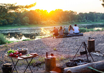 A company of young people sits on a log on the sandy bank of the river and looks at the sunset....
