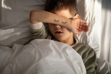 Tired peaceful caucasian little child sleeps on comfortable bed covers his face with hand with...