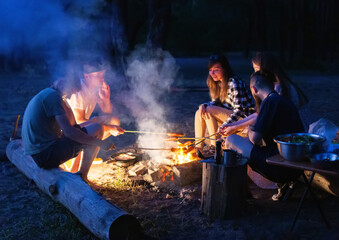 Young people are relaxing in nature in the evening, frying sausages on a fire and talking. Outdoor recreation in summer, happiness