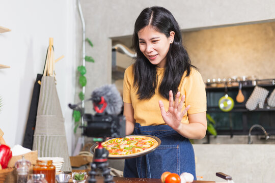 Young attractive Asian woman talking communicating during online live streaming how to cook homemade pizza indoor kitchen. content creator using camera recording food product review. - Powered by Adobe