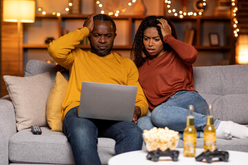 Shocked black spouses looking at laptop screen, home interior