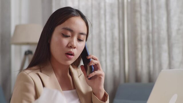 Close Up Of Asian Businesswoman Holding A Thermometer And Talking On Phone While Working With Laptop In The Living Room At Home
