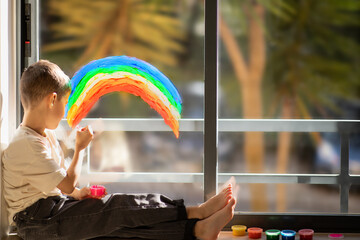Busy caucasian little child sits on floor with paints, draws rainbow on panoramic window in...