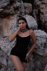 Obraz na płótnie Canvas Beautiful brunette in a black dress posing near the rocks and the sea. Attractive brunette woman in a little black dress on the sea coast.