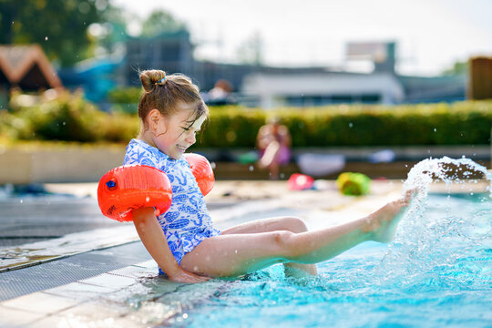 Little preschool girl with protective swimmies playing in outdoor swimming pool by sunset. Child learning to swim in outdoor pool, splashing with water, laughing and having fun. Family vacations.
