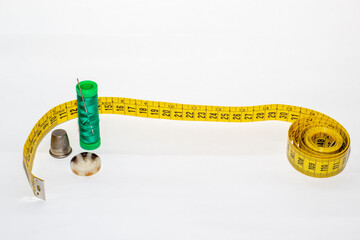 Bobina de hilo verde con un alfiler clavado junto un metro amarillo, un botón marrón y un dedal de metal sobre un fondo blanco. - Powered by Adobe
