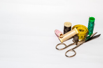 Bobinas de hilo de diferentes colores con un alfiler clavado junto una tijeras de coser, un metro amarillo, un botón marrón y un dedal de metal sobre un fondo blanco.