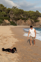 Hombre joven de raza caucásica jugando con su perro negro en la arena de la playa de la costa...