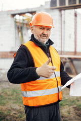 Senior man engineer or construction worker in hardhat approving building new modern house with blueprints. Portrait of male architect or contractor holding plans at construction site