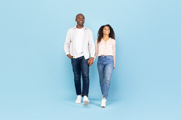 Happy african american couple walking, holding hands and looking away on blue studio background, full length