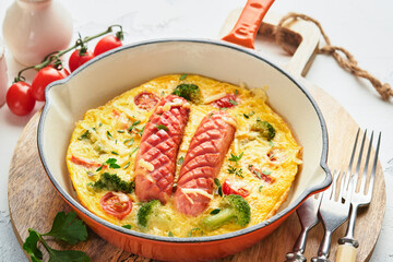 Omelette or frittata with two sausages, melted cheese, cherry tomatoes, broccoli and parsley on red skillet or iron pan on white old concrete background. Healthy breakfast food concept. Top view.