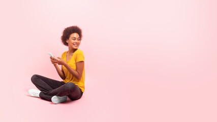 Happy young african american curly lady sit on floor, look back at empty space isolated on pink...