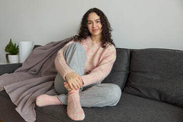 Portrait of a young curly smiling woman wearing cozy home clothes  sitting on the grey couch with...