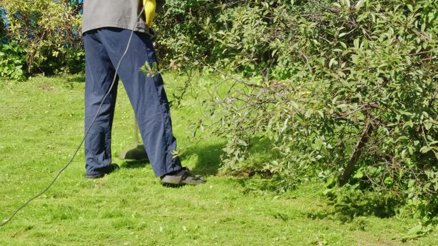 Faceless man mowing lawn with brush cutter. Back view. Leg, feet of male yardman in waterproof outdoor slippers who holds lawnmower. Gardener working on grass plot, backyard with grass trimmer