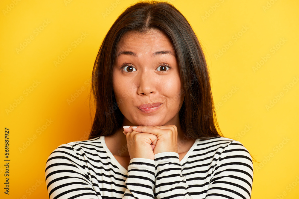 Wall mural Portrait shot of cute Asian female model keeps hands under chin presses lips smiles gently has dreamy expression looks to the camera wears striped pullover poses over yellow background - Wall murals