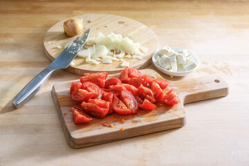 Sliced tomatoes, chopped onions, a knife and feta cheese on cutting boards and on a wooden kitchen counter, preparation of a healthy vegetarian salad, copy space, selected focus