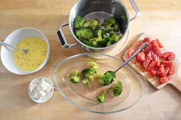 Broccoli in a pot, sliced tomatoes and onions, feta cheese and an egg mixture are filled into a glass baking dish, cooking preparation for a vegetable casserole, high angle view